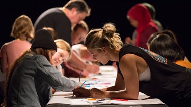 A group of children and adults colour on table covered by white paper.