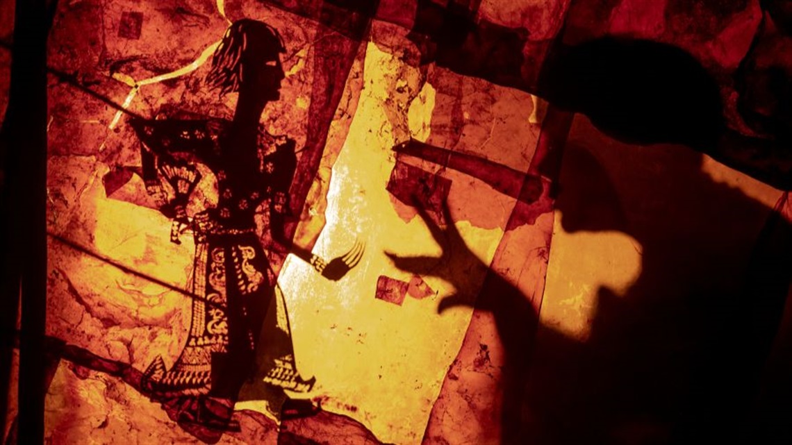 A burlap skin canvas projecting two shadowy dark puppets. The one on the left is a smaller than the puppet on the right.