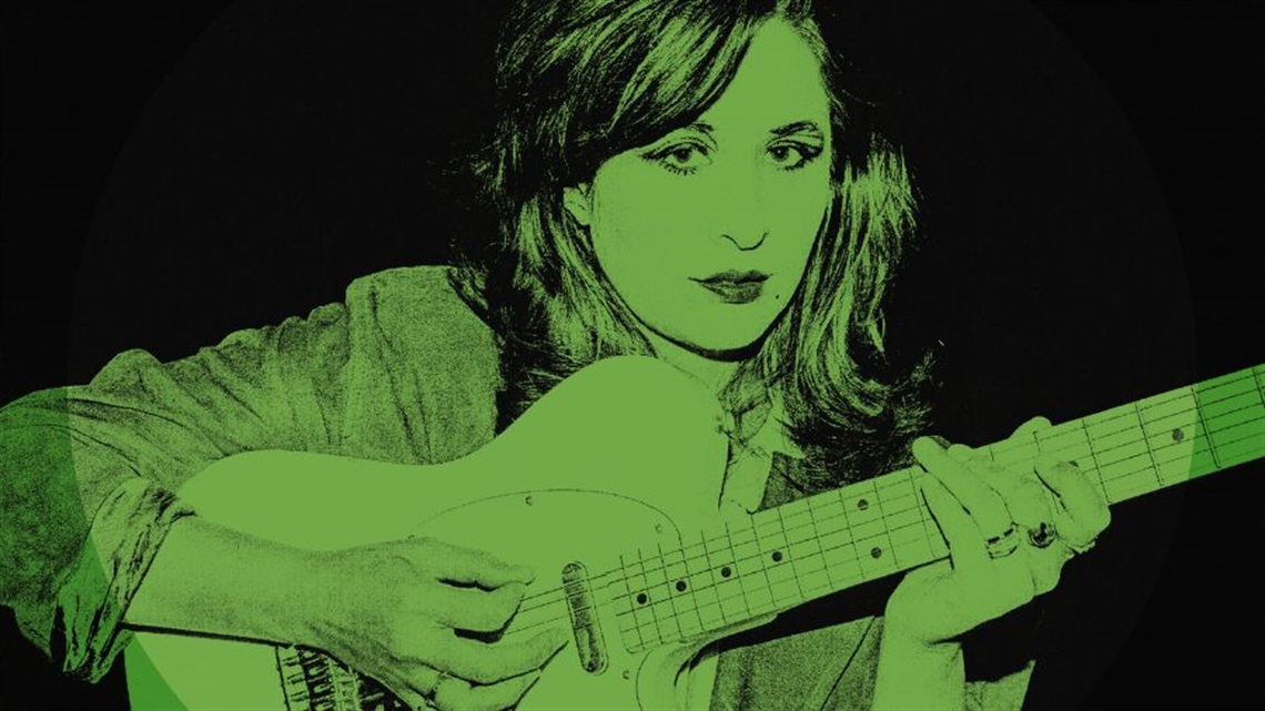 A woman with long hair holds a guitar in front of herself and looks sombrely to the camera. A green hue is over the image.