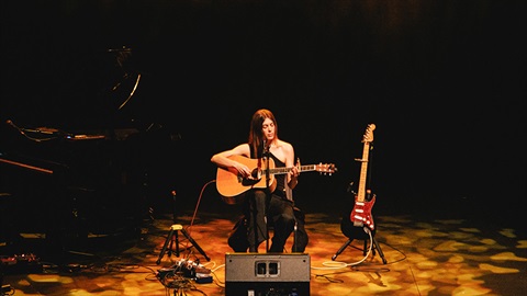 A woman with long dark hair sits behind a microphone on a dark stage. She holds an acoustic guitar on her lap. 