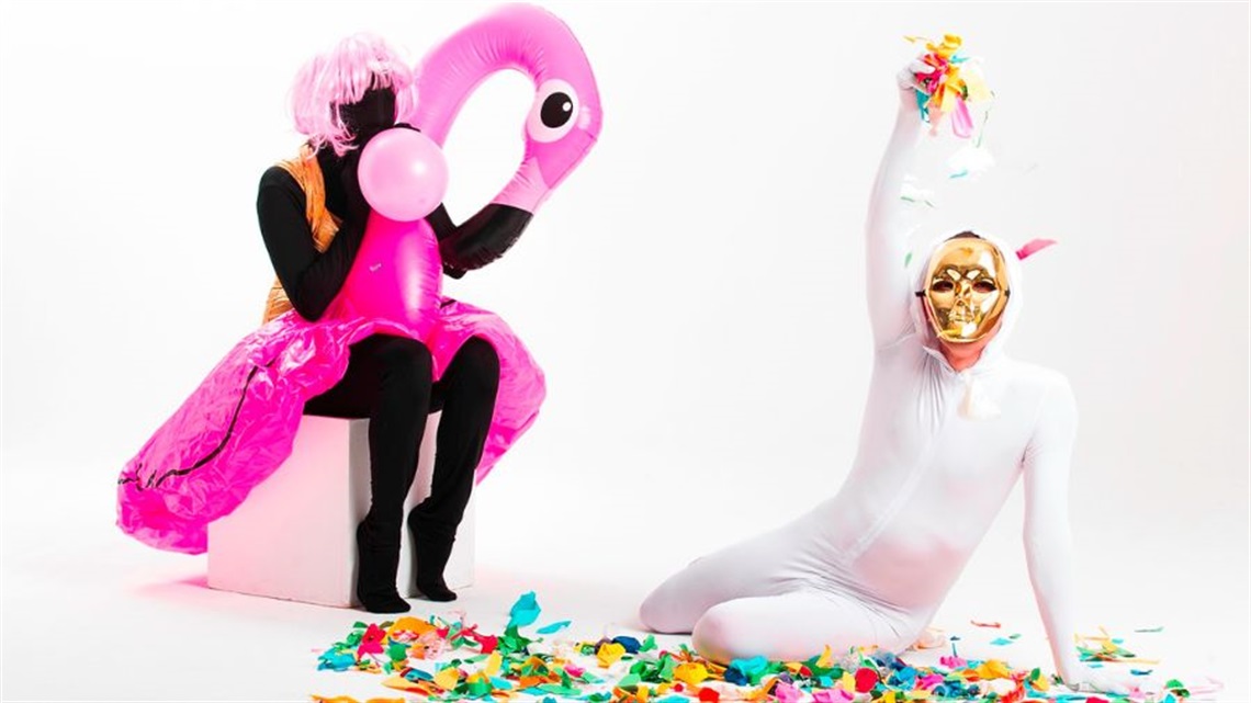 A person wearing a black full-body suit sits on a white box blowing up a pink balloon. Around their waist is a blow up flamingo. Another person on the right wears a full-body white suit and sits on a pile of confetti.