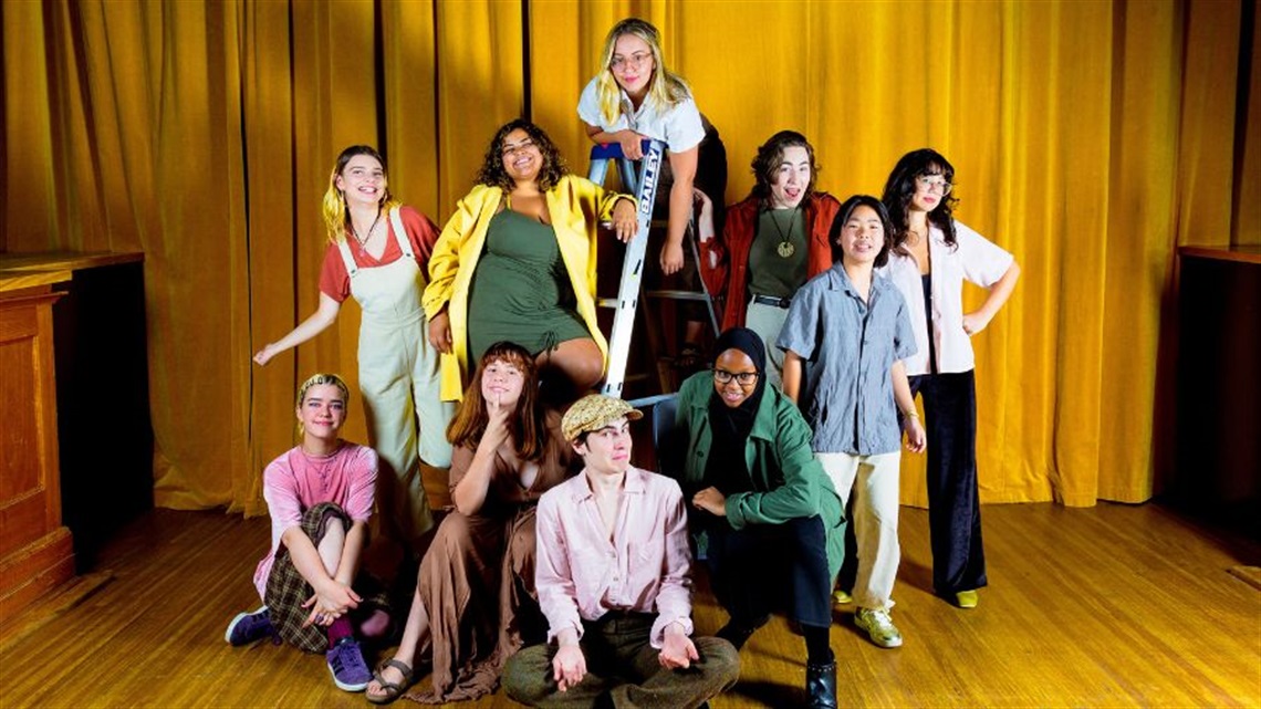 A group of young people gather around a ladder; some stand, some lean against the ladder, some sit on the floor in front. Behind them is a yellow curtain.