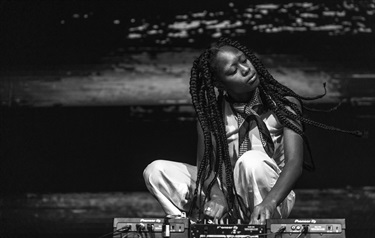 A black and white photo of a woman with long black braided hair crouches down to the floor with a music mixer in front.