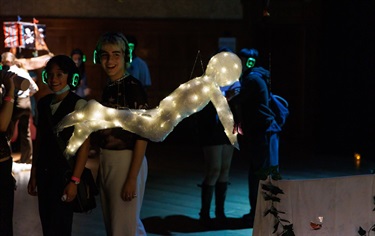 Two people look at hanging art work of a lit-up figure hanging from the ceiling with its arms and legs raised to the air.