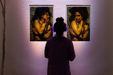 A silhouette of a person with a hair bun on their head stands with their back to the camera as they look towards two hanging pictures of framed art.