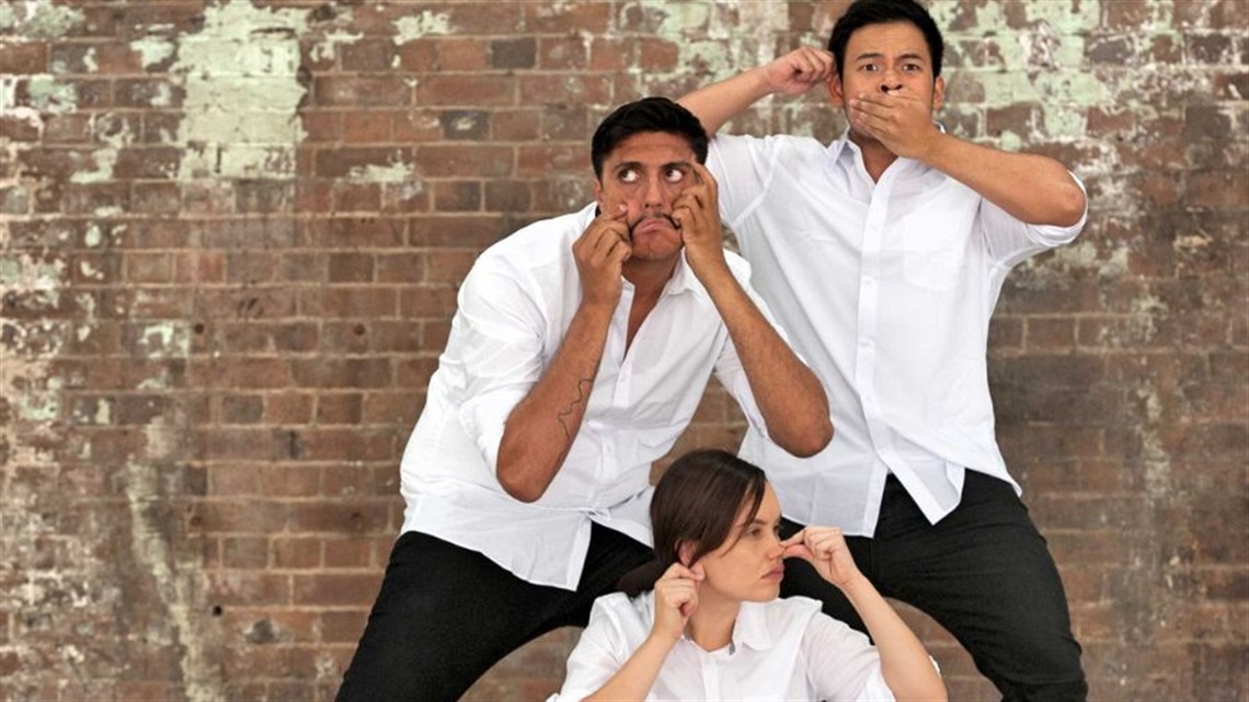 Two Aboriginal mena nd one Aboriginal woman wear white collared shirts and dark jeans. They cluster together in front of a brick wall with their hands touching their own faces.