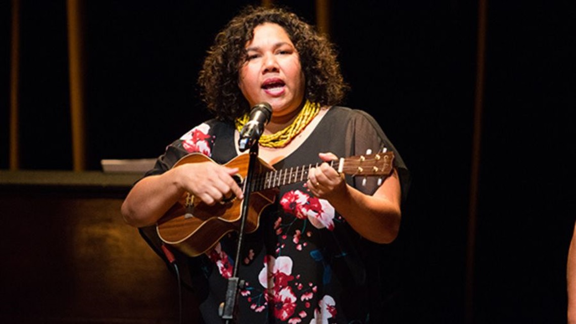 An Aboriginal woman with short curly dark hair is wearing a floral print black dress and a yellow necklace. She holds a ukele close to her chest in her ight hand and strums in with her left. She is singing into a microphone standing in front of her. 