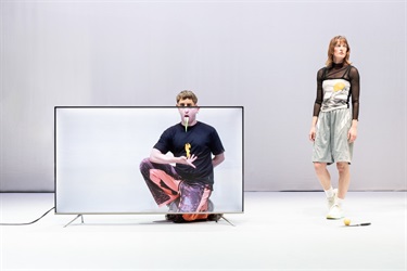 A man with short light hair and wearing a black tshirt perches behind a large screen tv. A woman with short light hair wearing a black sheer long sleeve top and pale green pants stands to the right.
