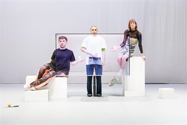 Three people stand behind a large tv screen. Two of them sit on white platforms while one person in the middle stands. Half of their bodies stick out from the tv screen while the other half projects on the tv screen.