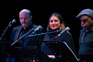 Three performers stand behind microphones