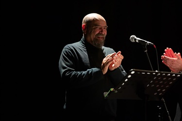 A man with a bald head stands smiling and clapping his hands behind a music stand.