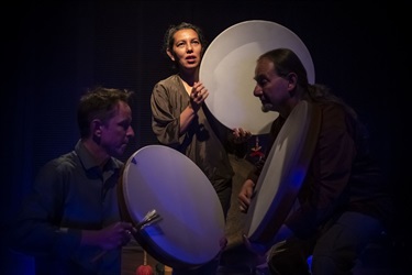 A woman holds a large drum head stands at centre stage. Crouched below and to the side of her are two men holding small instruments in their hands.