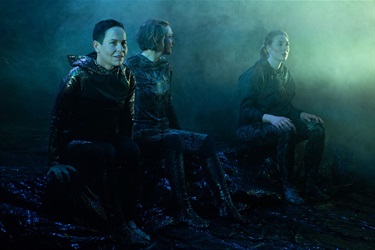 Three people wearing dark outfits and sitting on black objects surrounded by haze lit in green.