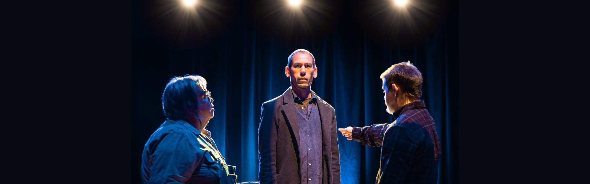 A bald-headed man wears a brown business jacket and buttoned shirt stands in the center under three spot lights. Two people surround him, with the person on the right pointing their right arm at him.