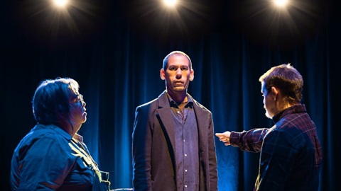 A man with thin light hair wears a dark suit. He s surrounded by two other people on either side of him and face towards him. The person on the right holds out their right arm.