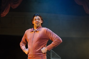 A man with brown hair is wearing a pink jumpsuit. His arms are perched on his hips in a superman pose.