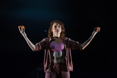 A woman with long curly hair and wearing a pink jumpsuit, exposing her white crop top underneath outstretches her arms to the side. In her hands are two apples.