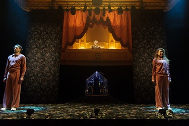 A large lit stage including a Gothic-like balcony has three people standing on it: a woman on the left side wearing a pink jumpsuit, a man with a large tall hat is in the balcony, centre stage and another man with dark hair wearing a jumpsuit.