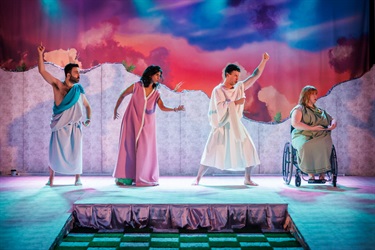 Two men and two women stand in a line across a raised white platform. They are frozen in various robotic-like positions with arms raised in the air. They wear toga outfits in various colours. The woman on the far right is sitting in a wheelchair.