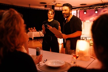A man wearing a black top and beige trousers stands in front of a white table set for diner service. He is holding a clipboard and smiling to the people seated at the table in front of him. A woman holding a camera is standing to his left.