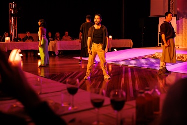 Four people are standing with their legs splayed in a box formation across a floor. A row of long white tables are behind and in front of the group.