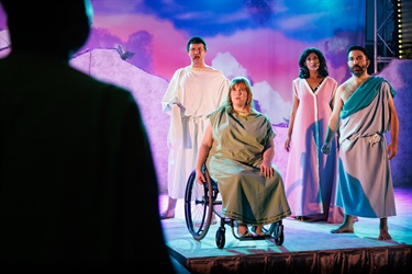 Two men and two women are clustered together, looking toward a dark shadow to the camera's left. They all wear toga outfits in various colours. One woman is in a wheelchair.
