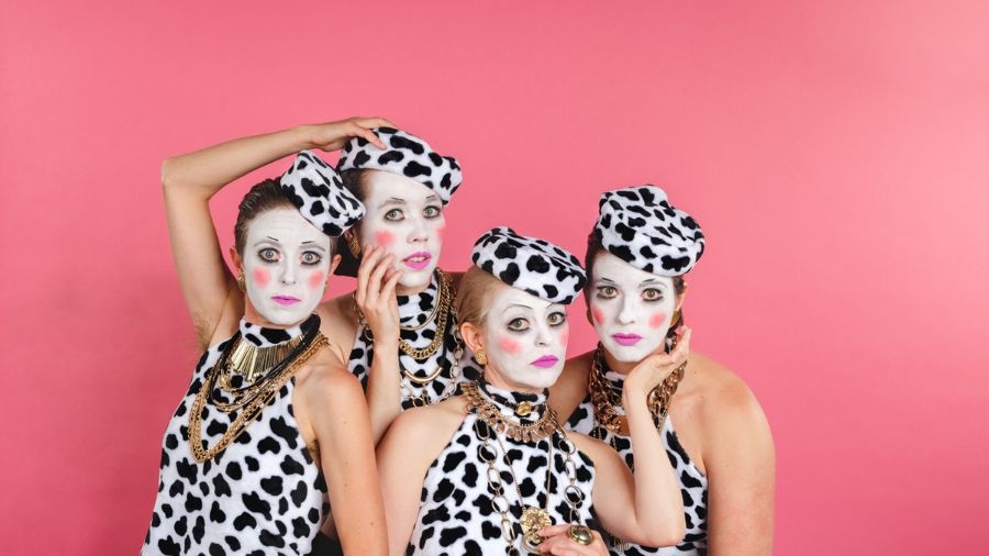 Four people wearing black and white cow patch tank tops and hats and gold-chain necklaces gather together in a cluster. Their faces are painted in white with black eyeliner outlining their eyes, pink on their cheeks and neon-pink lips.