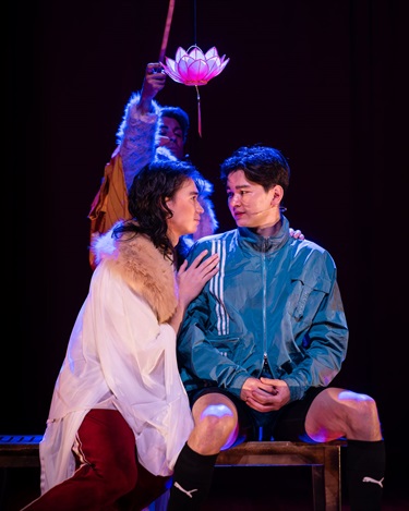 Two people sit next to one another. The person on the left is in profile, holding onto the shoulder of the other, looking longingly in the other's eyes. Above them is a lotus on a string. 