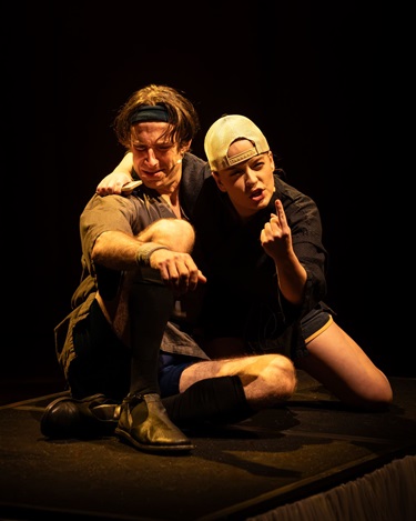 Two people in a warm glow crouch down on the floor. One of them wears a backwards cap and points their finger to make a point.