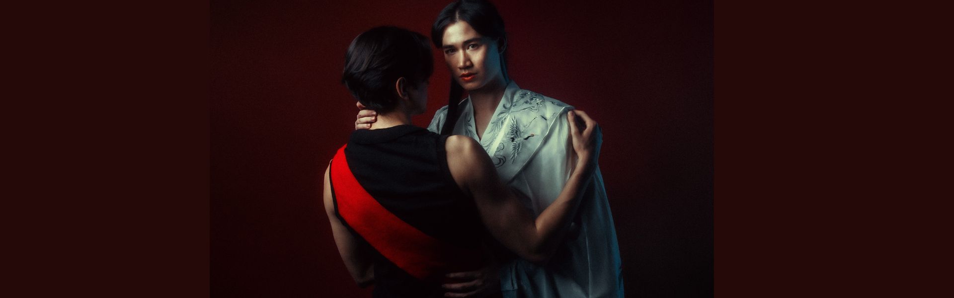 Two Asian people, one wearing a black sleeve-less top with a red diagonal stripe and with their back to the camera, is embraced by the other person wearing a white robe.