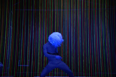 A person poses in front of a rainbow curtain with their face covered by blue hair.