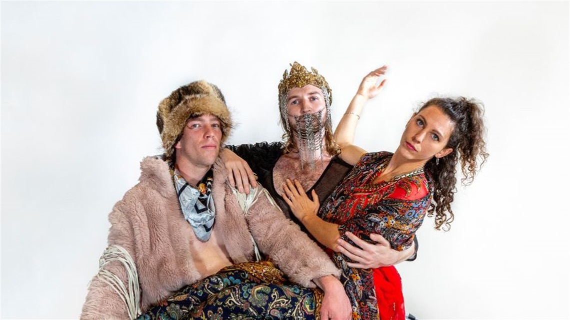 A woman with long curly hair sits across the lap of two men. She wears a red print dress and holds her right hand in the air with her elbow resting on a shoulder. The two men wear eclectic regal clothes. 