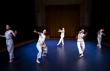 Five women stand across a black stage in various poses. They all wear white pants and white tops.