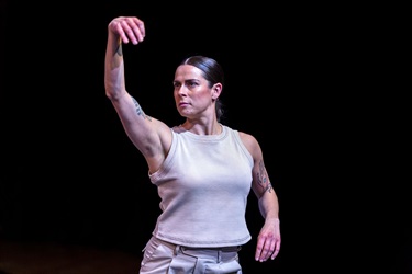 A woman with dark hair pulled back stands facing to the left. Her right arm is out stretched. She is wearing a white top.