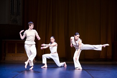 Three woman all wearing white pants and tops stand across a black stage in various poses.