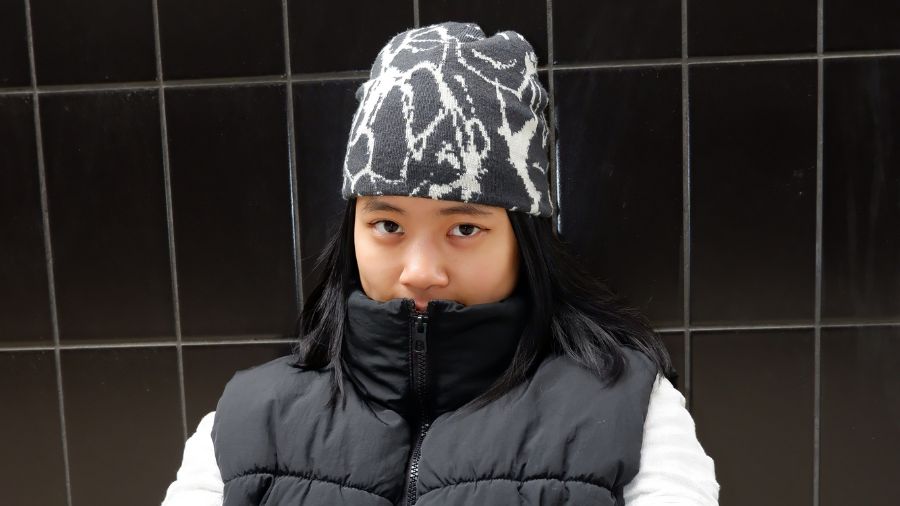 A person with long dark hair and wearing a black and white beanie on their head also wears a dark puffer vest and white long sleeve top. They stand in front of a black tilted wall.