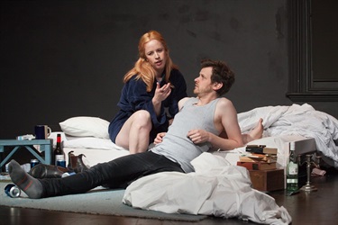 A man and a woman sit on a dishevelled bed, casually engaged in conversation with one another.