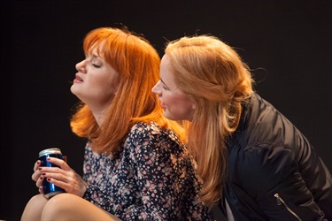 A woman with long light hair looks over the shoulder of a with red hair holding a can. 