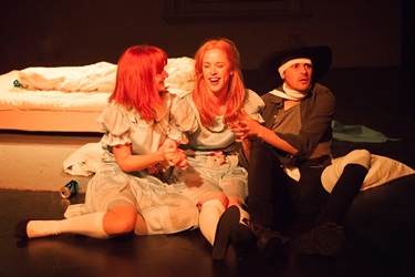 Two women and a man dressed in costumes sit side by side one another on a dark floor.