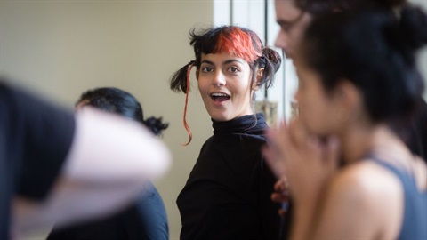 A person with dark, short hair and a streak of orange wears a black turtleneck shirt. They look towards the camera in surprise. Around them are blurred profiles of other people. 