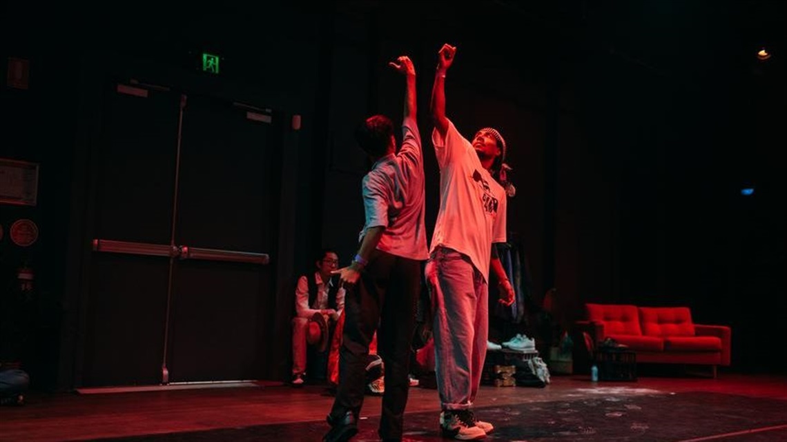 On a darkly lit stage, two people wearing loose clothing, stand next to one another, touching outstretched arms of each other and looking up.