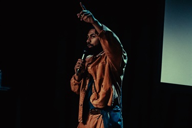 A man with a dark beard wears a brown jacket and trousers. He raises his right arm in the air and holds a microphone in his left to speak into it.
