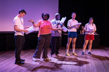 A group of people holding scripts stand across a stage.