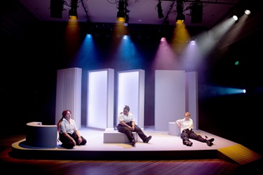 Three women wearing white security outfits sit across a lit stage. sitting on set.