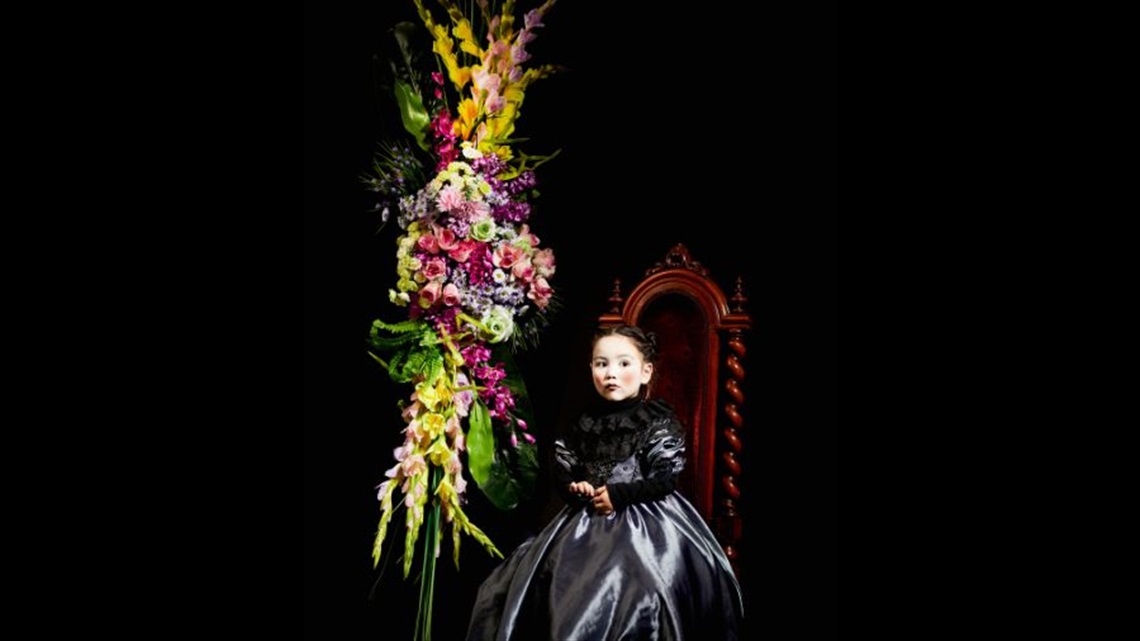 A young girl wearing a grey and black Victorian dress sits on a tall-back chair. Next to her is a tall bouquet of flowers.