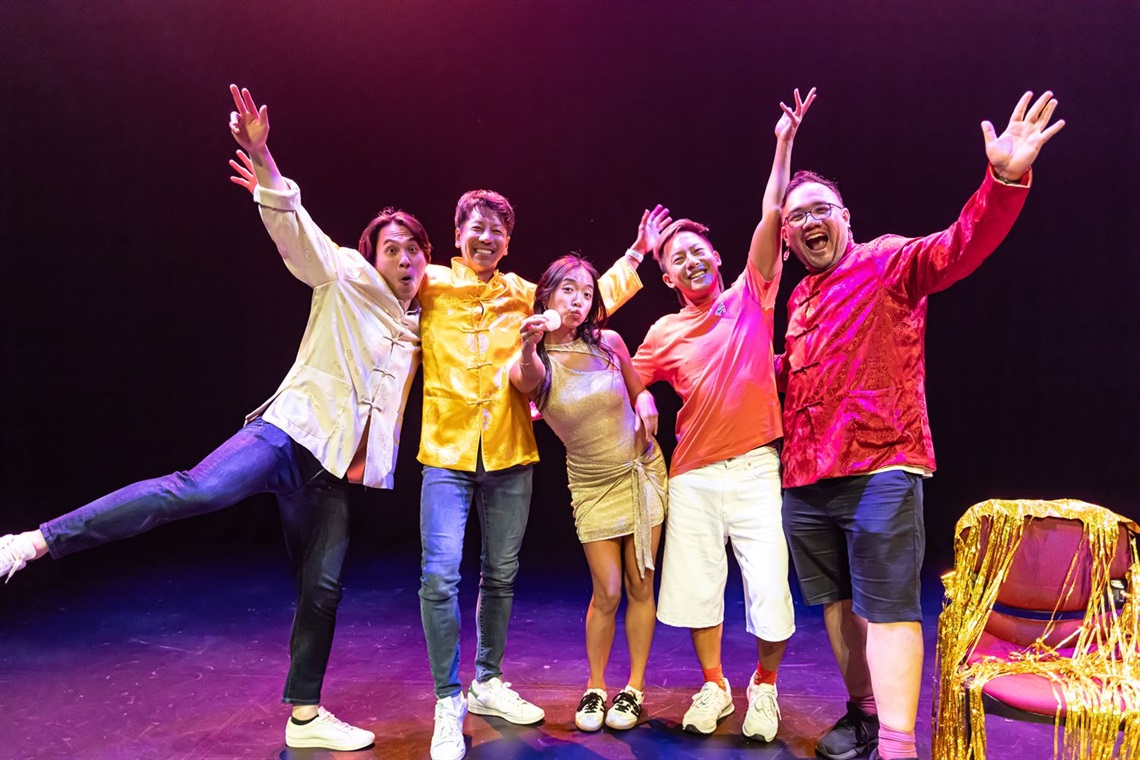 A group of four men and one woman, all of Asian descent, stand arm in arm next to each other a lit theatre stage. They raise their hands in the air in joy and celebration.