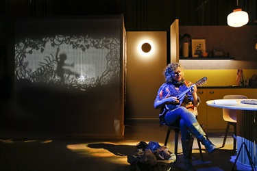 A woman strumming a guitar at a kitchen table. A screen projecting shadow puppets.