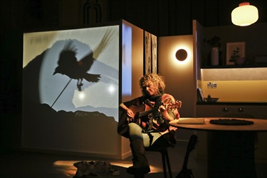 A woman plays the guitar at a round table, while the shaow puppet of a bird projects over her right shoulder.