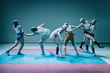 Six people wearing white tank tops, various coloured wrestling helmets and matching coloured shorts stand in a line across the stage. They move in various directions and positions.