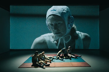 People sit on a mat in front a projection screen. The screen displays a person wearing a white wrestling helmet and white tank top.